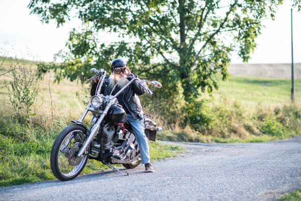 A senior man traveller with motorbike and goggles on road in countryside. Copy space.