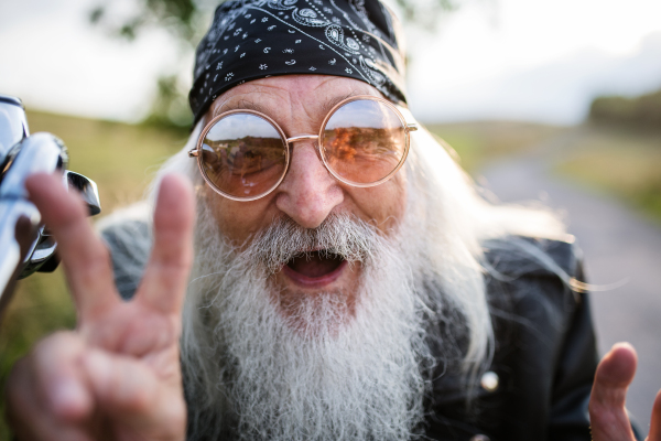 Close-up of senior man traveller with motorbike and sunglasses in countryside, having fun.