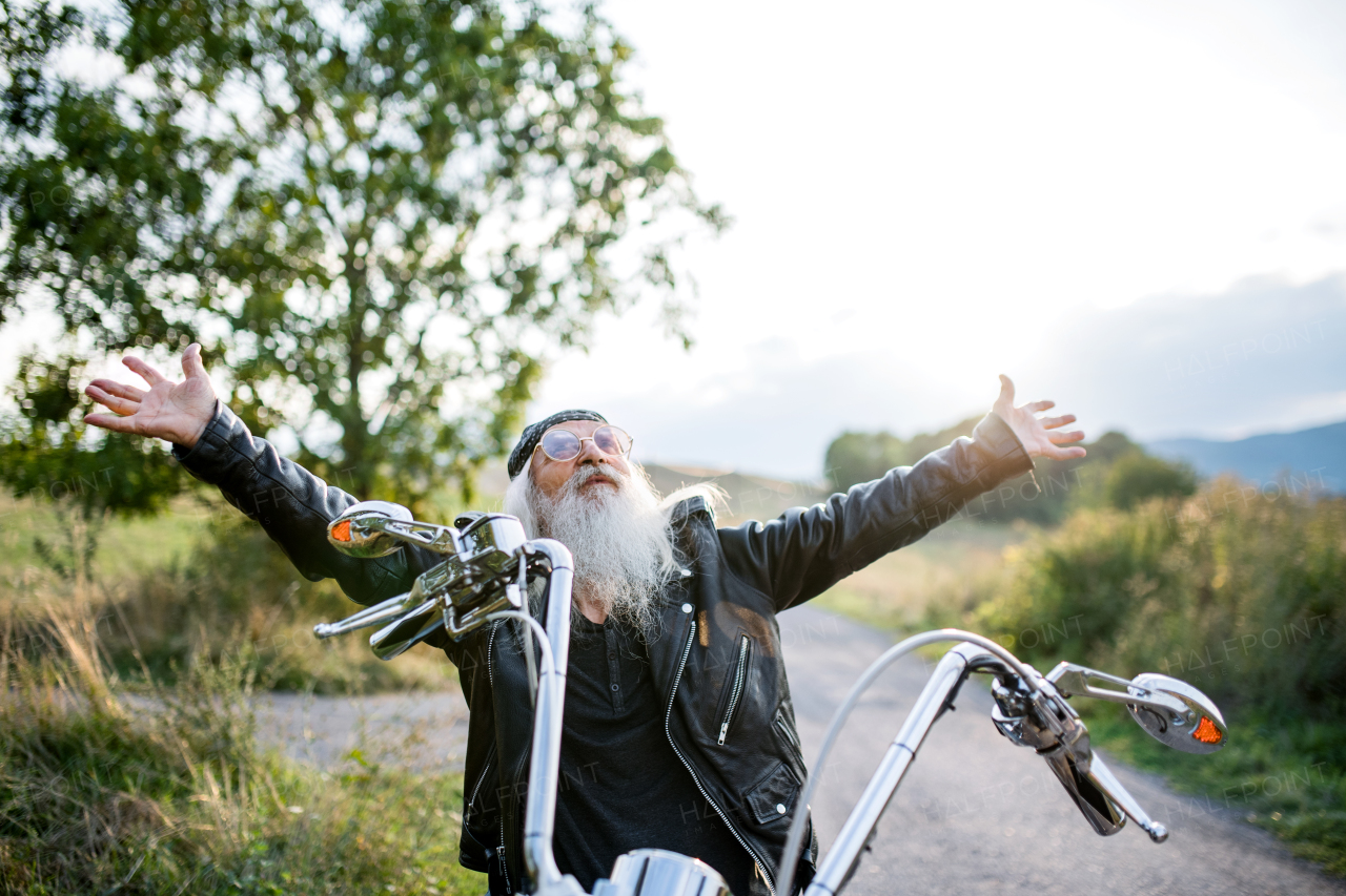 A senior man traveller with motorbike and sunglasses in countryside, stretching arms.