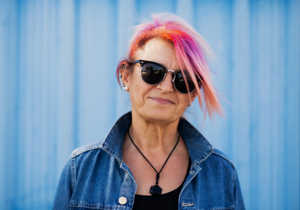 Portrait of senior woman with purple pink hair and sunglasses standing against blue background.