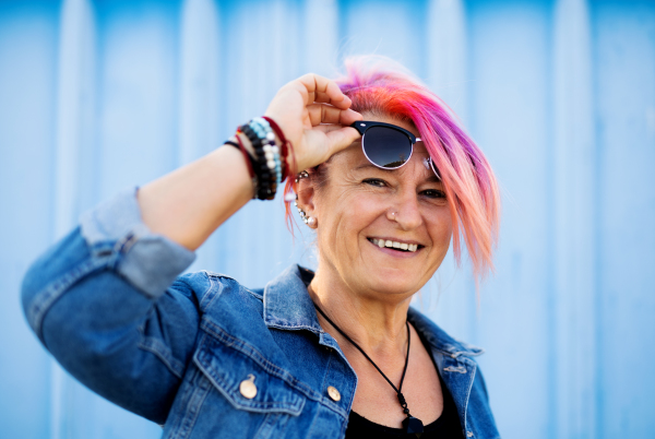 Portrait of senior woman with purple pink hair and sunglasses standing against blue background.