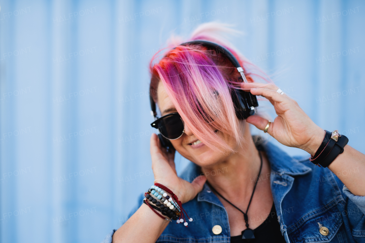 Portrait of senior woman with purple pink hair and sunglasses standing against blue background.