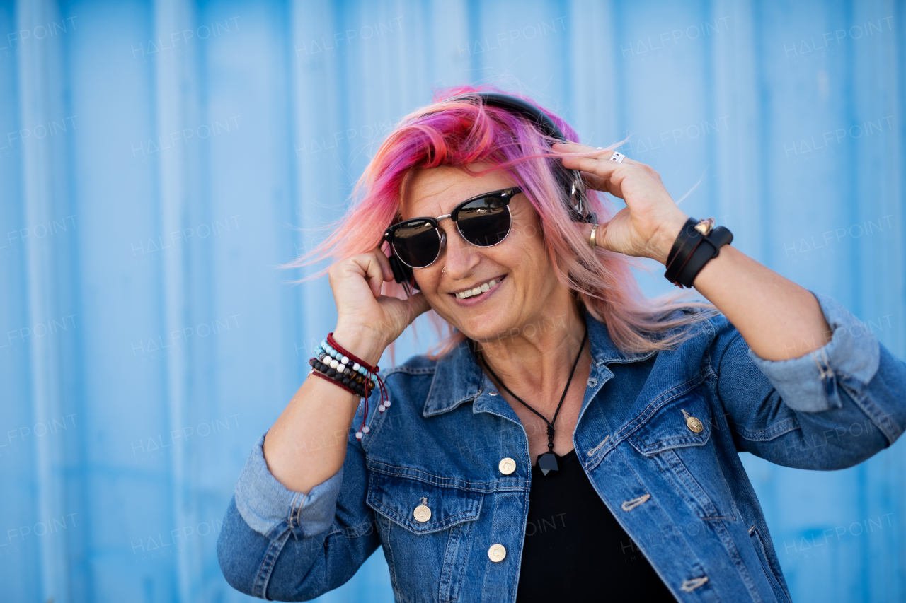 Portrait of senior woman with purple pink hair and sunglasses standing against blue background.