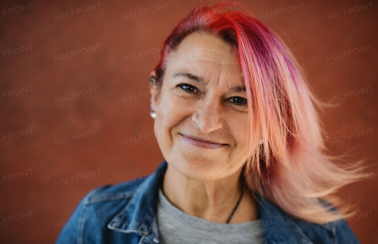 Portrait of senior woman with purple pink hair standing against brown background.