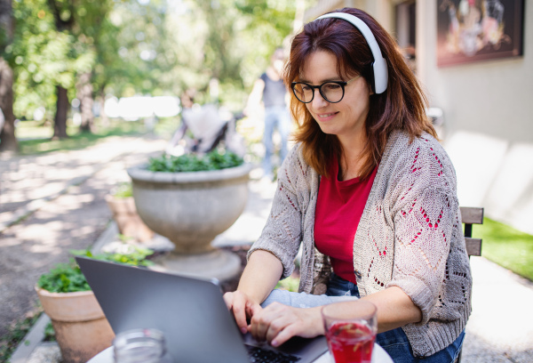 Mature woman with laptop and headphones outdoors in city or town park, working. Copy space.