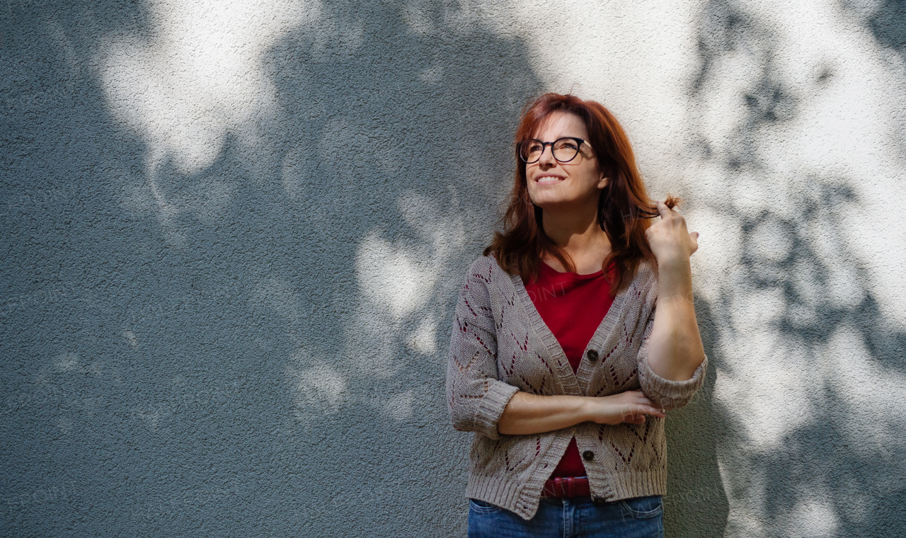 Portrait of mature woman standing outdoors in city or town against gray wall.