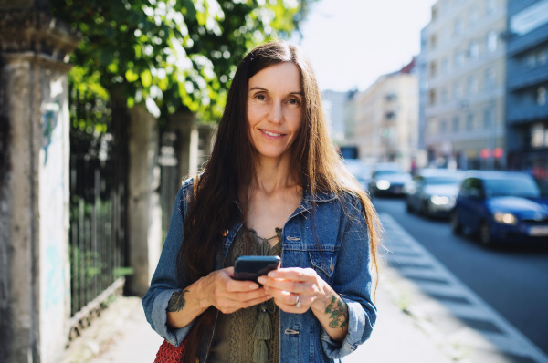 Portrait of mature woman with smartphone outdoors in city or town park, walking.