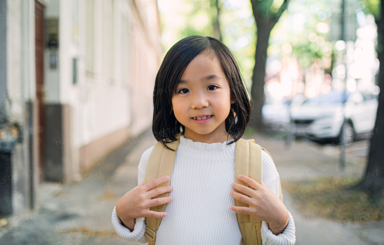 Portrait of small Japanese girl with backpack walking outdoors in town, looking at camera.