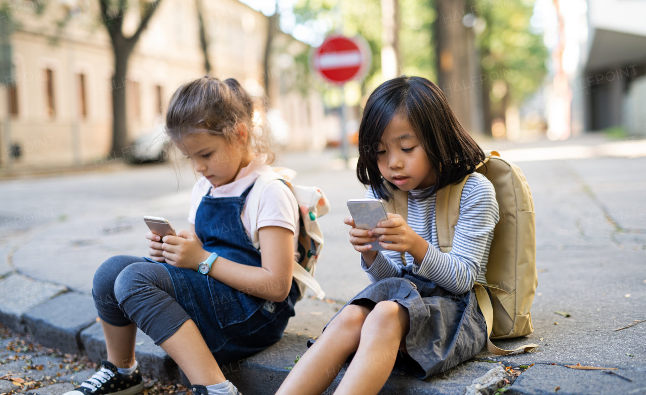 Small girls with smartphones sitting outdoors in town, playing online games.