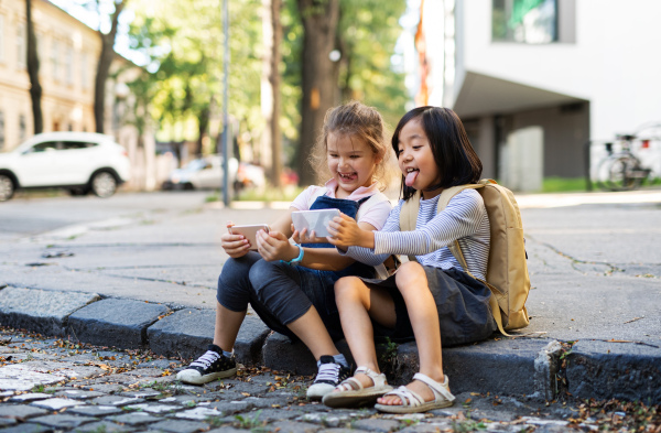 Small girls with smartphones sitting outdoors in town, taking selfie.