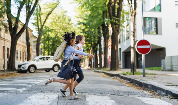 Side view of happy small school girls crossing street outdoors in town, running.