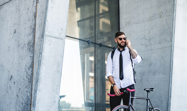 Portrait of young man commuter with bicycle standing outdoors in city, using smartphone.