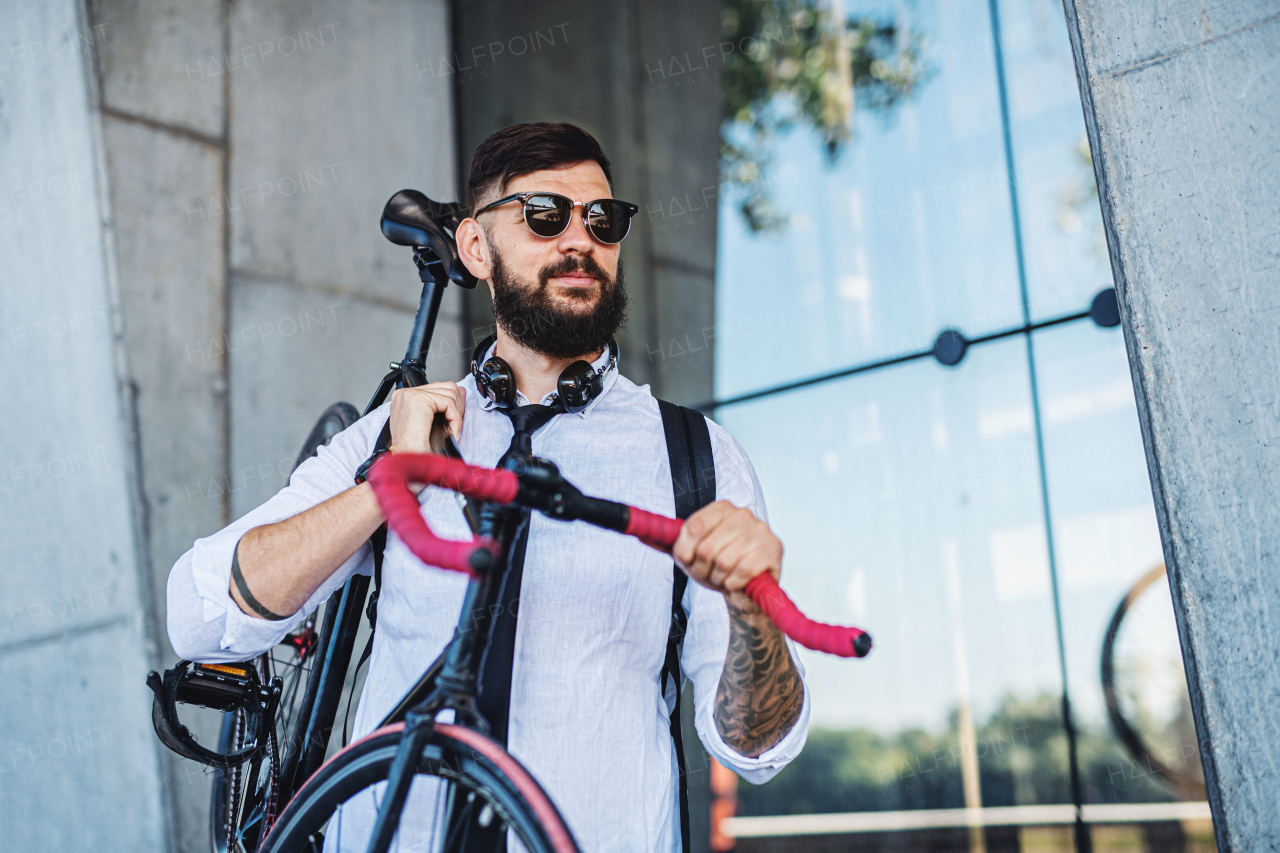 Portrait of young man commuter with headphones and bicycle walking outdoors in city.