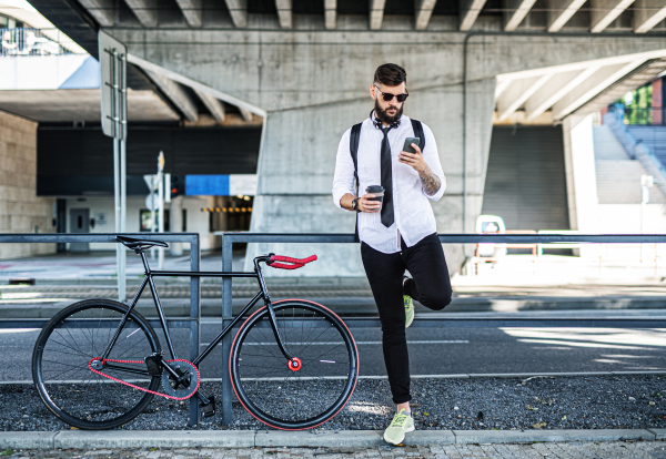 Portrait of young man commuter with bicycle standing outdoors in city, using smartphone.