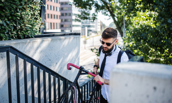 Portrait of young man commuter with headphones and bicycle walking outdoors in city.