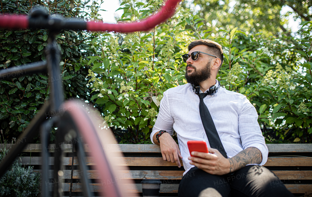 Portrait of young man commuter with bicycle sitting outdoors in city, using smartphone.