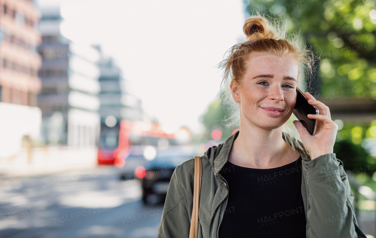 Front view portrait of cheerful young woman with red hair outdoors in town, using smartphone.