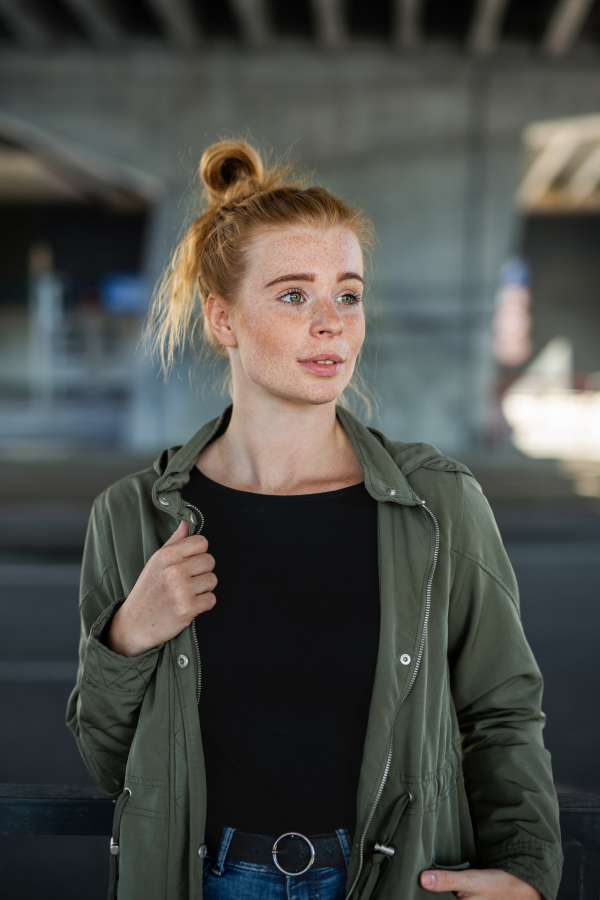 Front view portrait of cheerful young woman with red hair outdoors in town, standing.