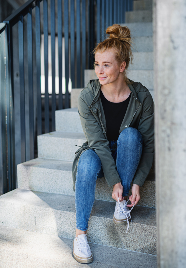 Portrait of young woman with red hair outdoors in town, sitting and tying shoelaces on staircase.