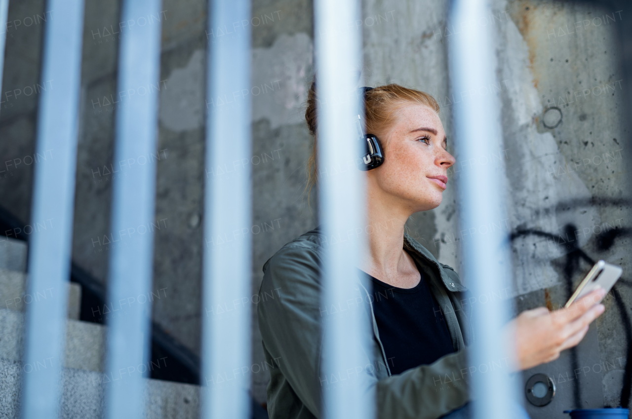 Portrait of young woman with red hair outdoors in town, using smartphone and headphones.