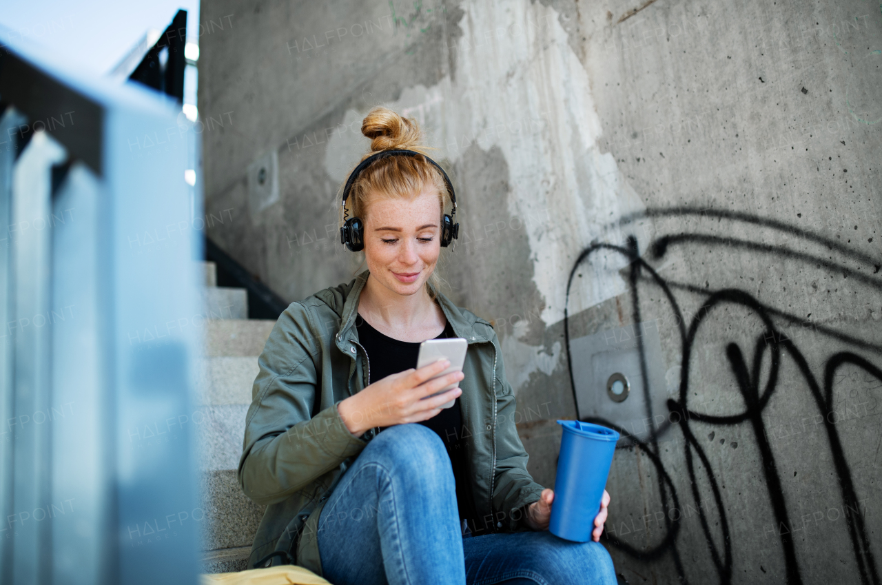 Portrait of young woman with red hair outdoors in town, using smartphone and headphones.