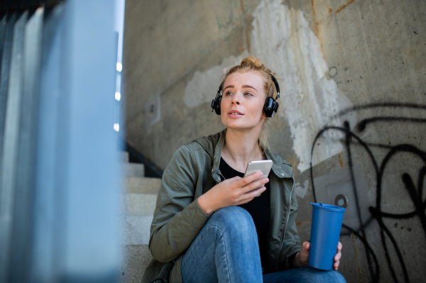 Portrait of young woman with red hair outdoors in town, using smartphone and headphones.