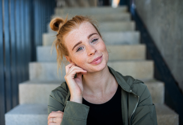 A front view portrait of young woman with red hair sitting outdoors in town, looking at camera.