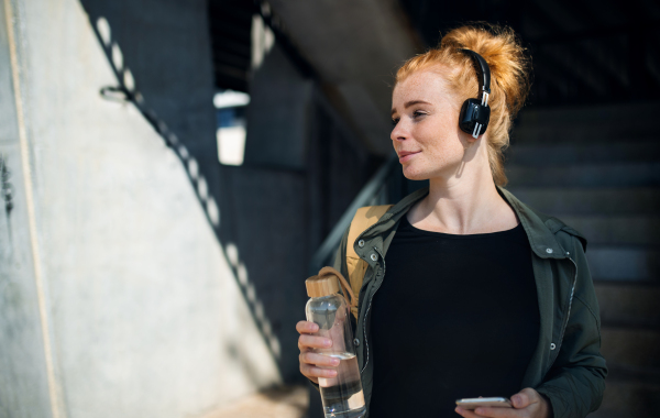 Portrait of young woman with red hair outdoors in town, using smartphone and headphones.