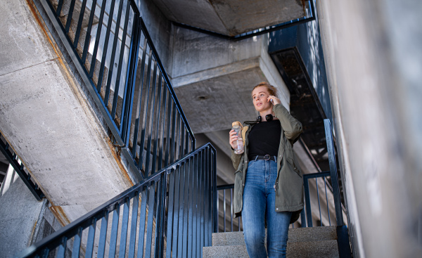 Portrait of young woman with red hair walking down stairs outdoors in town, using smartphone.