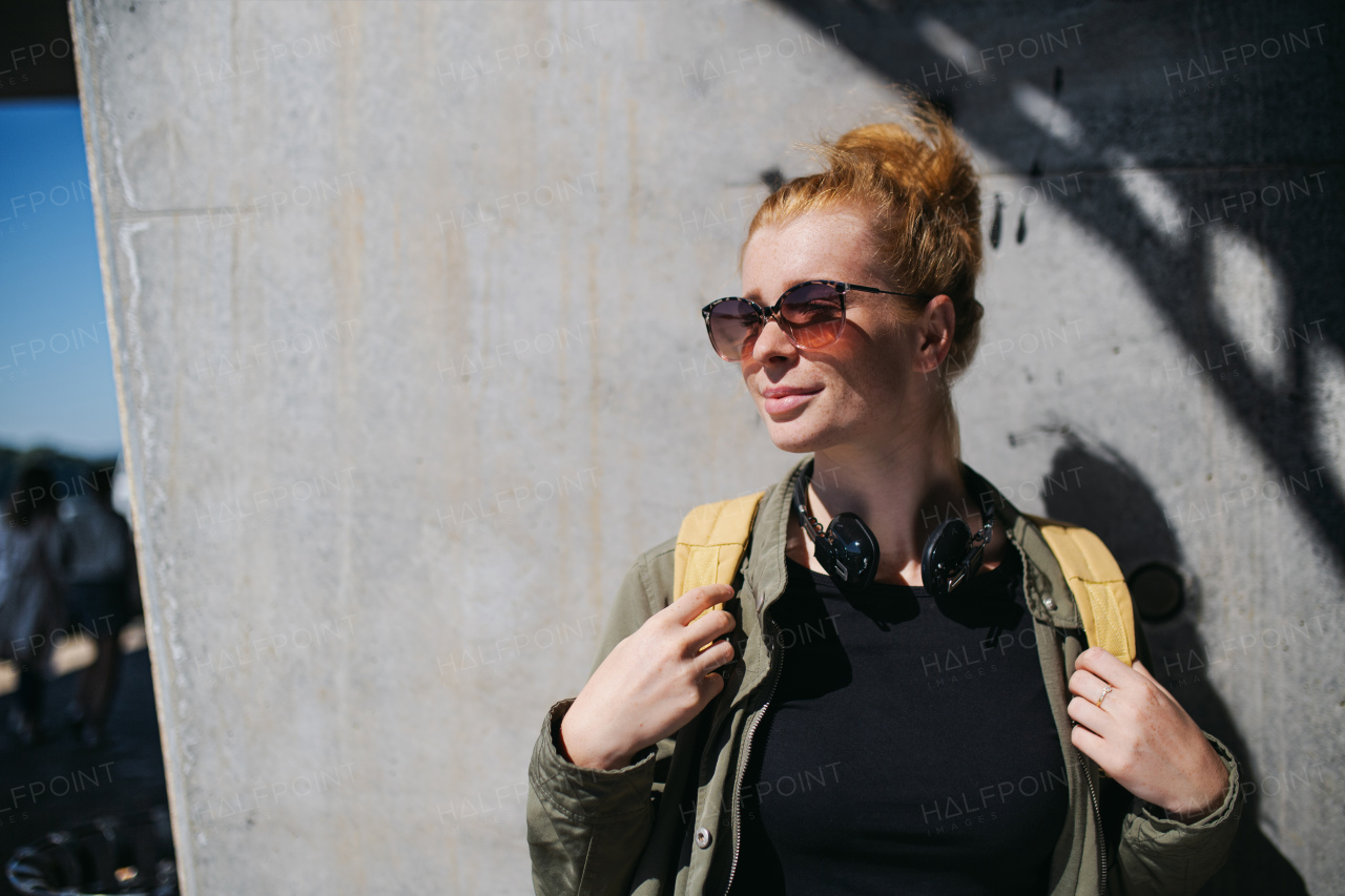 Front view portrait of cheerful young woman with red hair outdoors in town, wearing sunglasses.