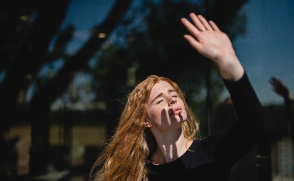 Portrait of young woman with red hair and closed eyes outdoors in town, blocking sunlight with hand.