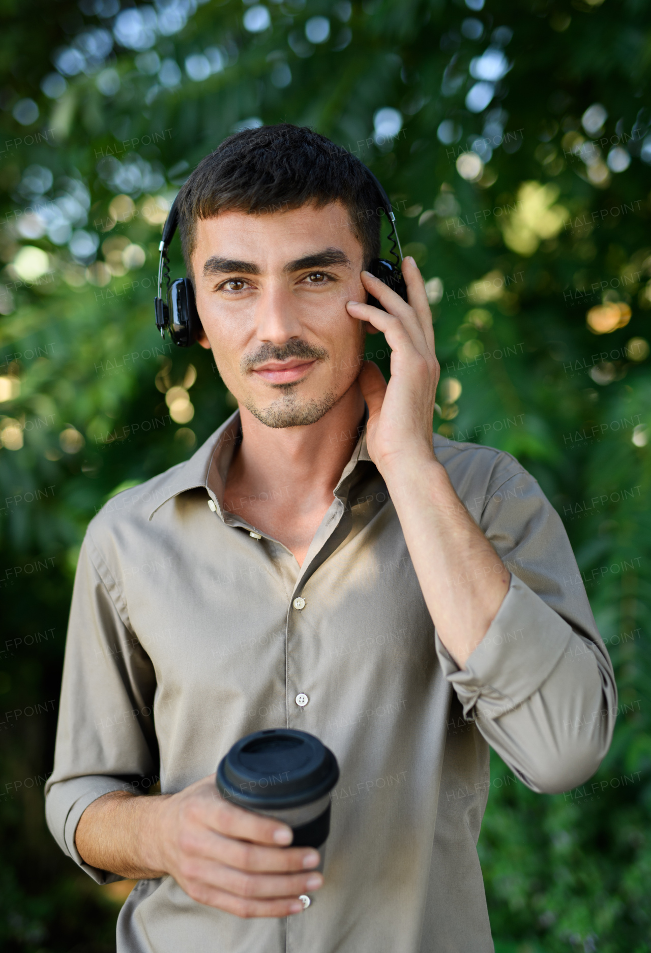 Young attractive man with headphones and coffee in reusable cup standing outdoors in city.