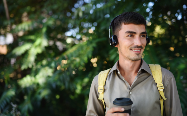 Young attractive man with headphones and coffee in reusable cup walking outdoors in city.