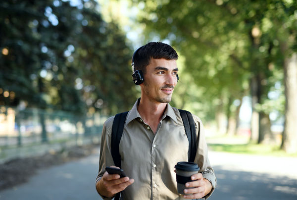 Young attractive man with headphones and coffee in reusable cup walking outdoors in city.