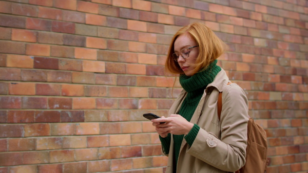 A mid adult woman using smartphone and walking against the brich wall in town.