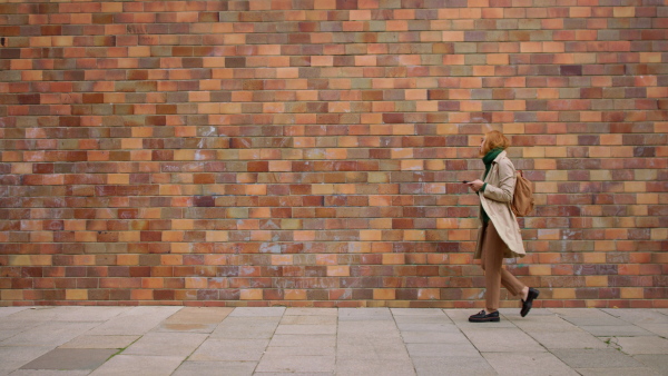 A mid adult woman using smartphone and walking against the brich wall in town.