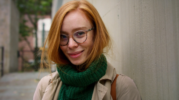 A mid adult woman using smartphone, waiting outdoors and leaning against concrete wall on autumn day, looking at camera.