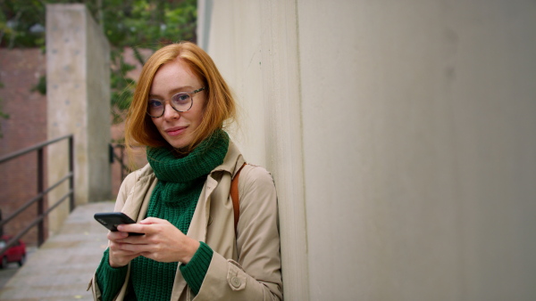 A mid adult woman using smartphone, waiting outdoors and leaning against concrete wall on autumn day, looking at camera.