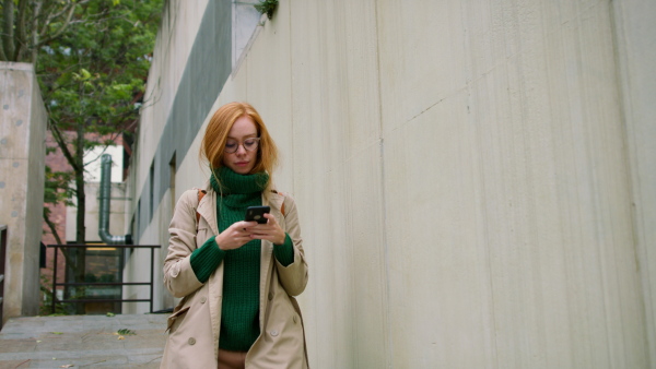 A young woman using smartphone and walking in street in autumn.