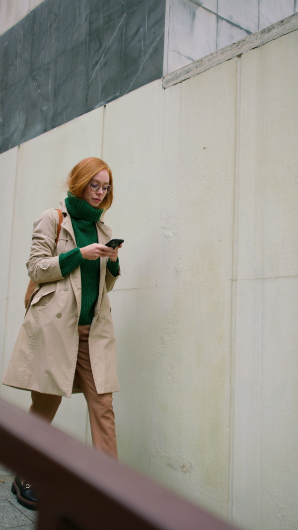 A vertical footage of young woman using smartphone and walking in street in autumn.