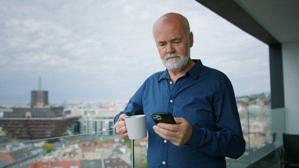 A senior man with coffee standing on balcony and making phone call at home.