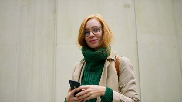 A mid adult woman using smartphone, waiting outdoors and leaning against concrete wall on autumn day, looking at camera.