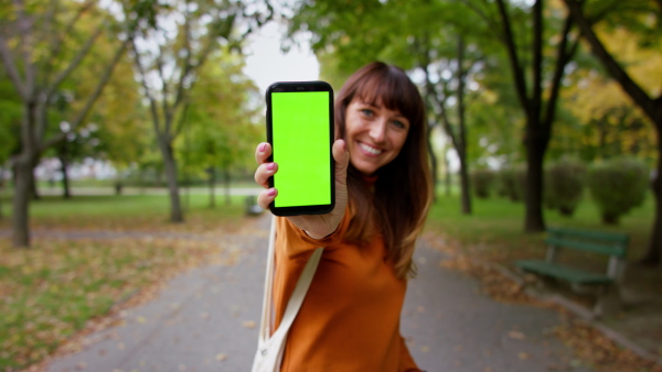 A beautiful mid adult woman showing smart phone looking at camera outside in park on autumn day.