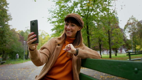 A beautiful mid adult woman sitting on bench and having video call in park in autumn.