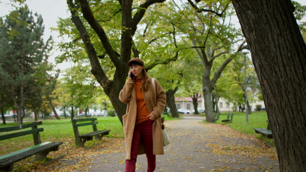 A mid adult woman in warm coat and knitted hat holding smart phone and walking in park.