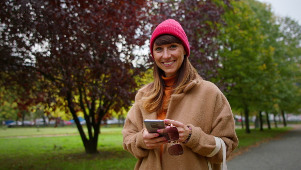 A mid adult woman in warm coat and knitted hat holding smart, laughing and looking at camera in park.