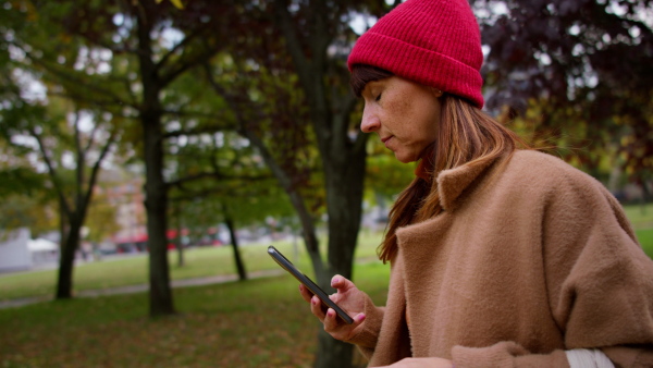 A mid adult woman in warm coat and knitted hat holding smart phone and walking in park.