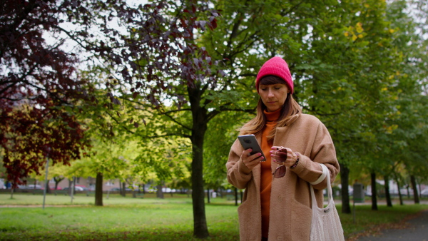 A mid adult woman in warm coat and knitted hat holding smart phone and walking in park.