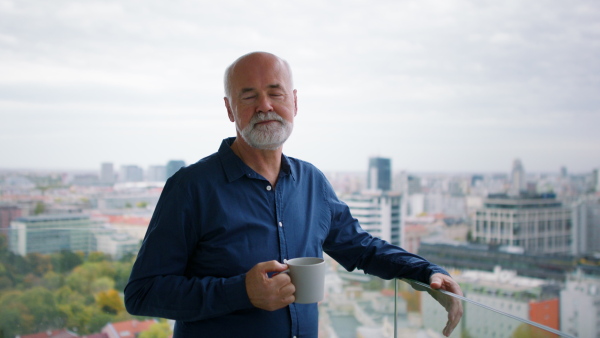 A senior man with coffee standing on balcony and looking at camera at home.