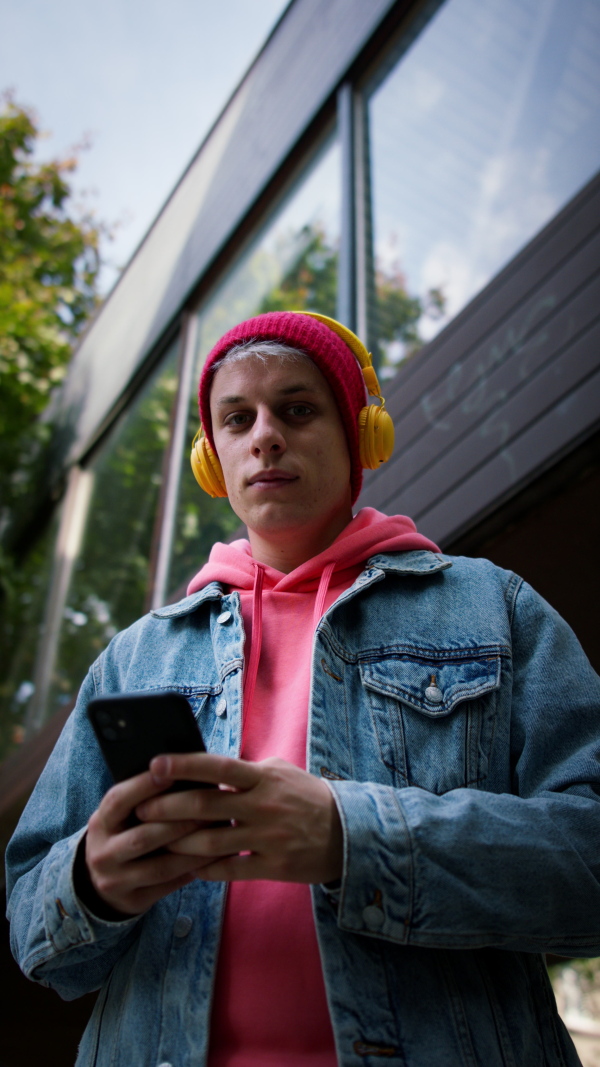 A low angle view of cool young man with headphones and smartphone outdoors in town, looking at camera.